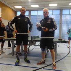 Zwei Männer in Sportkleidung halten einen Pokal in einer Tischtennishalle, während andere Spieler im Hintergrund applaudieren.
