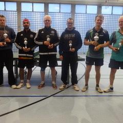 Sechs Tischtennisspieler stehen in einer Sporthalle und halten Pokale in der Hand. Helle, neutral gehaltene Umgebung.