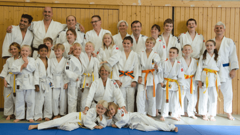 Gruppenfoto von Judoka unterschiedlichen Alters in weißen Judogis, lächelnd auf einer Trainingsfläche.