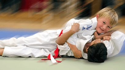 Zwei Kinder im Judogi kämpfen auf der Matte; eines liegt oben und lächelt, während das andere darunter liegt.