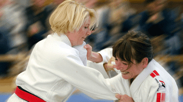 Zwei Frauen in Judogewändern engagieren sich in einem Wettkampf auf einer blauen Matte. Zuschauer im Hintergrund.