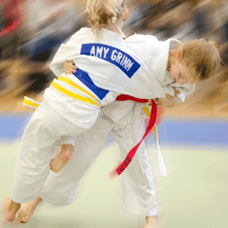 Zwei Kinder im Judoanzug üben einen Wurf; eines trägt einen roten, das andere einen gelben Gürtel.