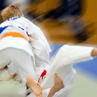 Zwei Kinder im Judoanzug kämpfen auf einer Matte, während einer den anderen zu Fall bringt. Bewegungsunschärfe sichtbar.
