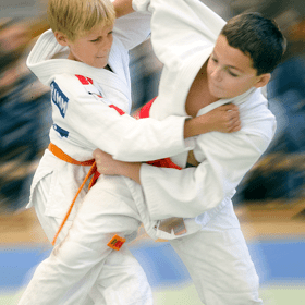 Zwei Kinder im Judogi zeigen einen Wettkampfbewegung in einer Sporthalle. Einer zieht am Anzug des anderen.