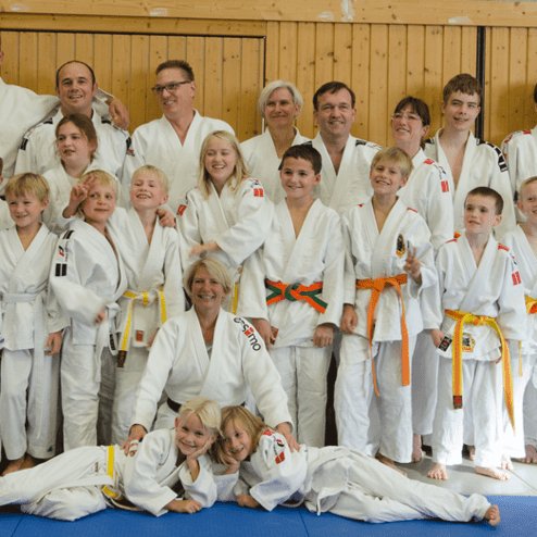 Gruppenfoto von Judoka unterschiedlichen Alters in weißen Judogis, lächelnd auf einer Trainingsfläche.