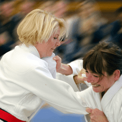 Zwei Frauen in Judogewändern engagieren sich in einem Wettkampf auf einer blauen Matte. Zuschauer im Hintergrund.