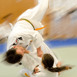 Zwei Judoka führen einen Wurf im Wettkampf aus, einer wird in die Luft gehoben. Die Bewegungen sind dynamisch verschwommen.