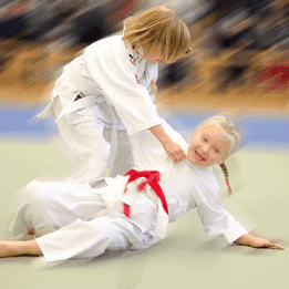 Zwei Kinder im Judogi kämpfen auf einer Matte, eines zieht das andere mit einem Griff zu Boden.