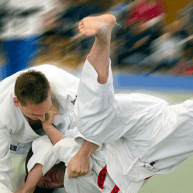 Zwei Judoka in Weiß kämpfen auf einer Matte, einer führt einen Wurf aus, während Zuschauer im Hintergrund aufmerksam zuschauen.