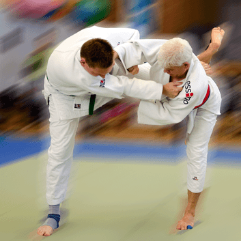 Zwei Judokas führen einen Wurf im Gelände aus, einer hebt den anderen an, beide tragen Judogis.