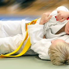 Zwei Kinder im Judoanzug kämpfen auf einer Matte, einer hat einen gelben Gürtel, der andere einen weißen.