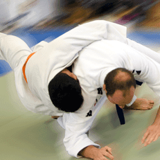 Zwei Judokas in weißen Gi kämpfen dynamisch auf einer Tatami, während einer den anderen zu Boden bringt.