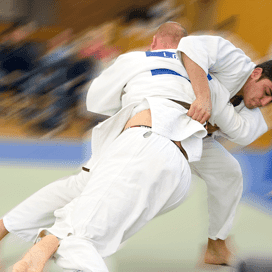 Zwei Judoka in Weiß und Blau kämpfen auf einer Matte, einer führt einen Wurf aus und der andere weicht aus.