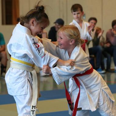 Zwei Kinder im Judoanzug k&auml;mpfen fr&ouml;hlich auf einer Judomatte, w&auml;hrend Zuschauer im Hintergrund applauds.