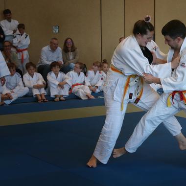 Judo-Training: Ein Mann und eine Frau &uuml;ben einen Wurfgriff, umringt von zuschauenden Kindern und Erwachsenen.