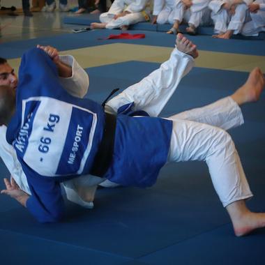 Zwei Judokas k&auml;mpfen auf einer blauen Matte, w&auml;hrend Zuschauer im Hintergrund sitzen und zuschauen.