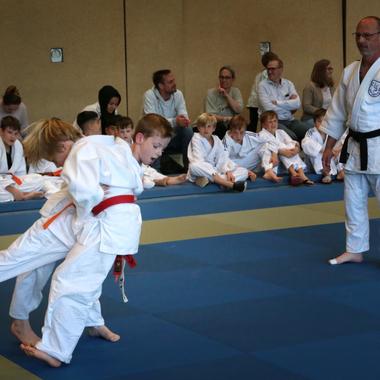 Zwei Kinder im Judoanzug &uuml;ben einen Wurf, w&auml;hrend ein Schwarzgurt-Lehrer zuschaut. Zuschauer sitzen am Rand der Matte.