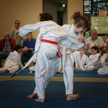 Zwei Kinder im Judoanzug k&auml;mpfen auf einer blauen Matte, Zuschauer applaudieren im Hintergrund.