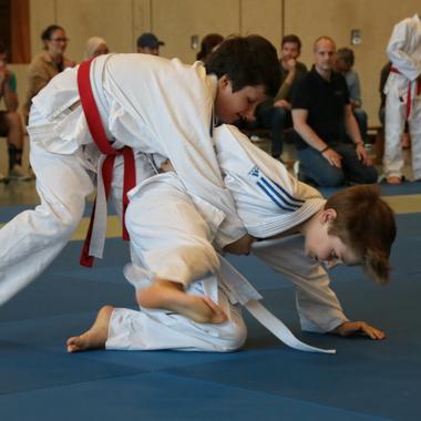 Zwei Kinder im Judoanzug k&auml;mpfen auf einer blauen Matte, w&auml;hrend Zuschauer am Rand zuschauen.