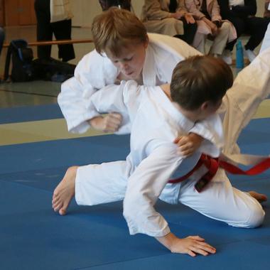 Zwei Jungen im Judoanzug k&auml;mpfen auf einer blauen Matte, einer greift den anderen an. Zuschauer im Hintergrund.