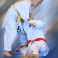 Zwei Kinder in weißen Judogi kämpfen; eines zieht das andere mit gelbem und rotem Gürtel zu Boden.