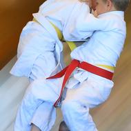 Zwei Kinder im Judoanzug führen einen Wurf im Training aus, einer trägt einen roten, der andere einen gelben Gürtel.