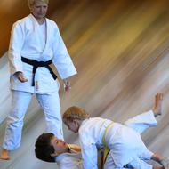 Drei Kinder im Judogi üben einen Kampf, während eine Trainerin ihnen zusieht. Bewegungseffekt im Hintergrund.