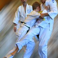 Zwei Kinder im Judogi üben einen Wurf, während ein Trainer im Hintergrund beobachtet. Dynamische Bewegung und Konzentration.