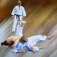 Zwei Judoka in weißen Anzügen führen eine Hebeltechnik aus, während ein dritter Beobachter zuschaut.