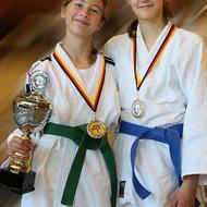 Zwei Mädchen in Judojacken zeigen stolz einen Pokal und Medaillen, eine mit grünen, die andere mit blauen Gürtel.