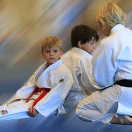 Zwei Kinder in weißen Judoanzügen mit einem Erwachsenen, der einen schwarzen Gürtel trägt, sitzen auf einer Judomatte.
