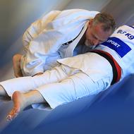 Zwei Judokas im Kampf, einer von hinten auf dem anderen liegend, beide in weißen Judoanzügen.