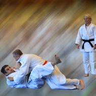 Zwei Judokas im Wettkampf, einer im Wurfansatz, der andere liegt am Boden; im Hintergrund eine dritte Person in Judokleidung.