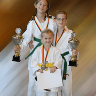 Drei Kinder in Judogi halten Pokale und Medaillen, stehen lächelnd vor einem verschwommenen Hintergrund.