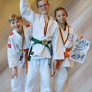 Drei Kinder in Judoanzügen stehen zusammen, eines hält einen Pokal, alle tragen Medaillen.