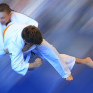 Zwei Judoka in weißen Anzügen führen einen Wurf auf einer blauen Matte aus, mit Bewegungsunschärfe im Hintergrund.
