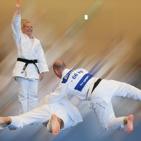 Judo-Wettkampf: Eine Kämpferin mit schwarzem Gürtel steht triumphierend, während zwei Männer im Wurf sind.