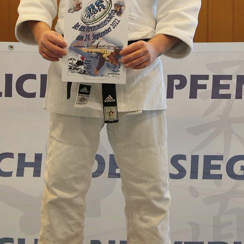 Mann in einem Judoanzug steht auf einem Siegertreppchen und hält eine Urkunde bei einer Sportveranstaltung.