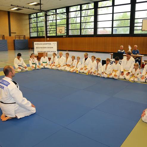 Gruppe von Judokas in weißen Gis, sitzend im Kreis auf einer blauen Matte, während sie Anweisungen erhalten.
