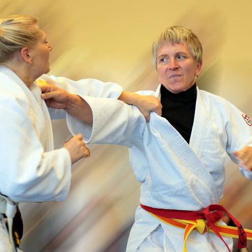 Zwei Judoka, eine mit blonden Haaren, üben eine Technik im Judogi in einer Trainingsumgebung.