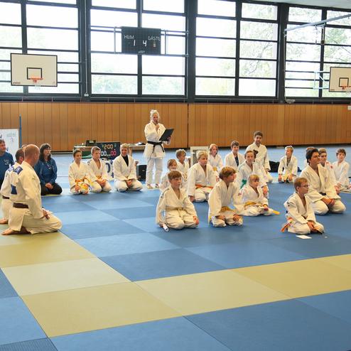 Gruppe von Judoka in weißen Anzügen sitzt auf einer tatami, während ein Leiter Anweisungen gibt. Im Hintergrund eine Uhr.