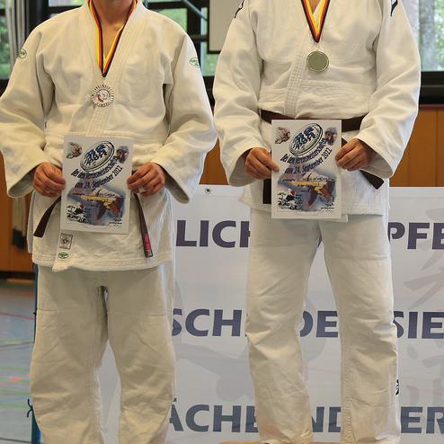 Zwei Judoka in weißen Anzügen stehen auf Podesten und halten Urkunden, im Hintergrund ein Banner mit „HERZLICH WILLKOMMEN“.