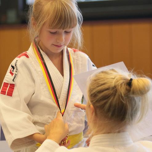 Ein Mädchen in einem Judoanzug erhält eine Medaille von einer Trainerin in einem Judo-Trainingsraum.