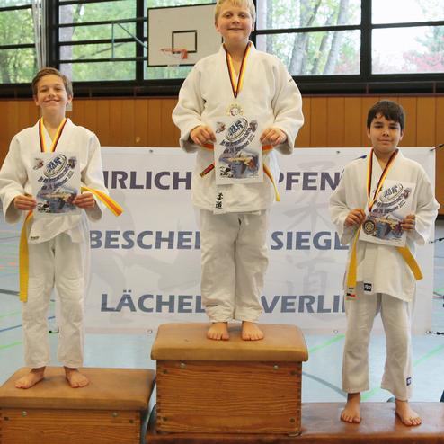 Drei Jungen in Judokleidung stehen auf Siegertreppchen und halten Urkunden bei einer Judo-Veranstaltung.