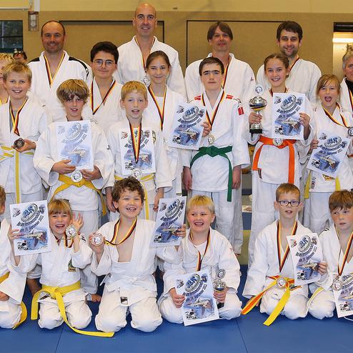 Gruppe junger Judoka in weißen Gi mit Medaillen und Urkunden, posierend auf einer Judomatte.
