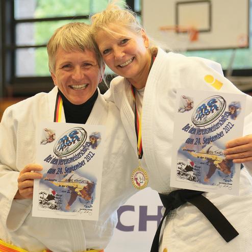 Zwei Frauen in Judoanzügen halten stolz Urkunden und eine Medaille nach einem Wettkampf. Hintergrund mit Fenster und Basketballkorb.