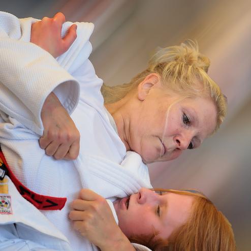 Zwei Judoka in weißen Gis kämpfen im Stand, die Trainerin fixiert die Schülerin am Oberteil.