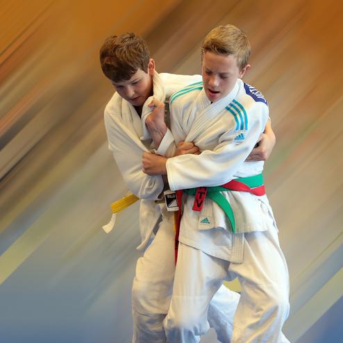 Zwei Jugendliche im Judoanzug üben einen Halt, während sich der Hintergrund farblich fast radial verwischt.