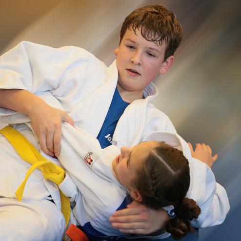 Zwei Kinder im Judoanzug kämpfen auf der Matte; eines hat das andere im Haltegriff.