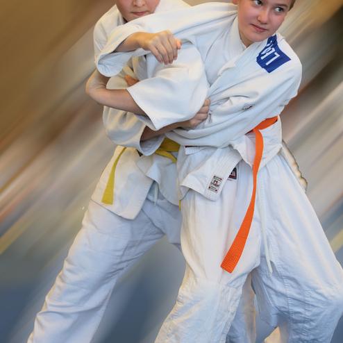 Zwei Kinder in Judoanzügen kämpfen in dynamischer Haltung auf einer judospezifischen Unterlage.